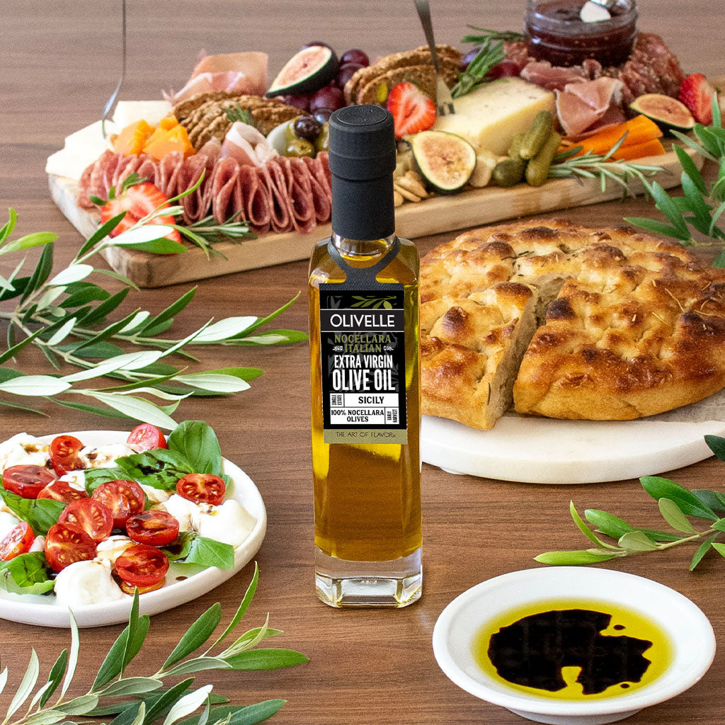 Olivelle olive oil bottle on a wooden table with food items including bread, salad, and cured meats.