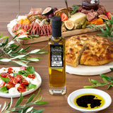 Olivelle olive oil bottle on a wooden table with food items including bread, salad, and cured meats.