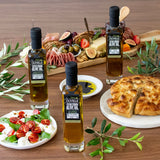 Three bottles of Olivelle olive oil on a wooden table with food items like bread, salad, and fruits.