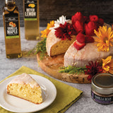 Vanilla cake with flowers on a wooden board, slice on a plate, and bottles of vanilla maple and vanilla bean extract in the background.