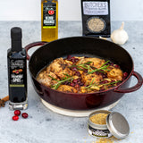 Dish of roasted chicken with cranberries and herbs, surrounded by cooking ingredients and spices on a gray surface.