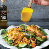 Salad with chicken and vegetables being drizzled with a yellow dressing, next to a bottle of Olivelle maple wood smoked bacon oil.