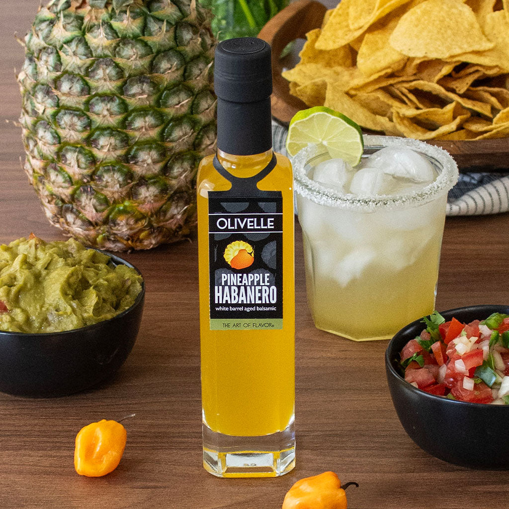 Olivelle Pineapple Habanero sauce bottle with a pineapple, chips, guacamole, and margarita on a wooden table.