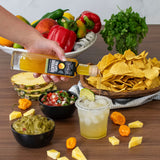 Hand pouring a bottle of pineapple juice over a glass with a wooden table full of snacks and fruits.