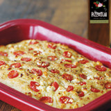 Rectangular pizza with cherry tomatoes in a red dish on a wooden surface, with a bottle of Pizzaiolo wine in the background.