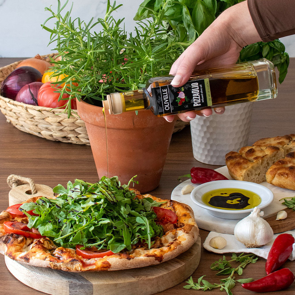 Person pouring olive oil over a pizza with greens on a wooden table with vegetables and bread.
