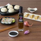 Bottle of Olivelle Roasted Shallot olive oil on a wooden table with sushi, dumplings, and shallots.