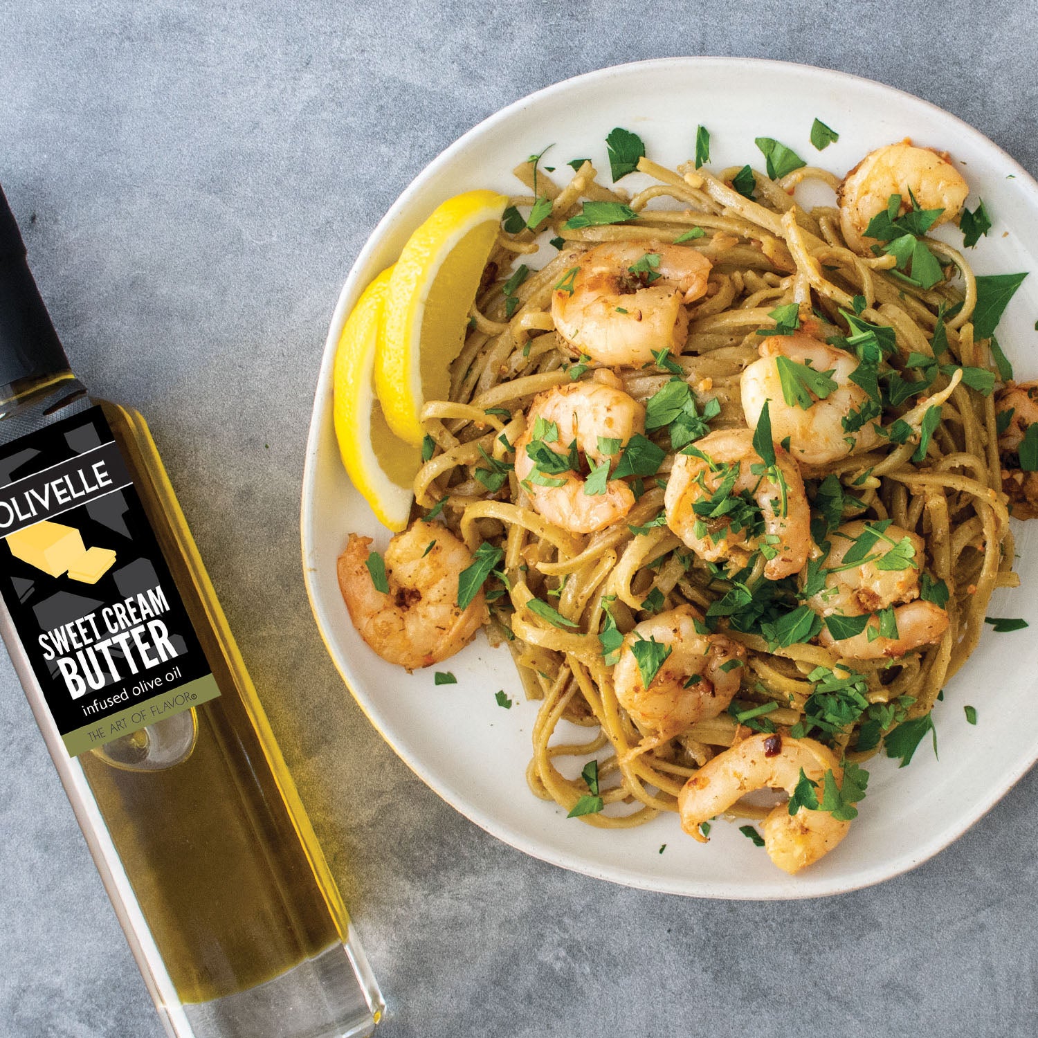 Plate of pasta with shrimp and lemon wedges, next to a bottle of Olivelle sweet cream butter on a gray surface.