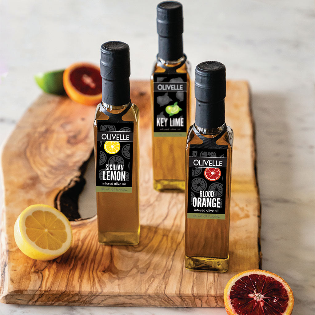 Three bottles of Olivelle olive oils on a wooden cutting board with citrus fruits.