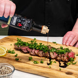 Person pouring sauce over a steak with herbs on a wooden cutting board.