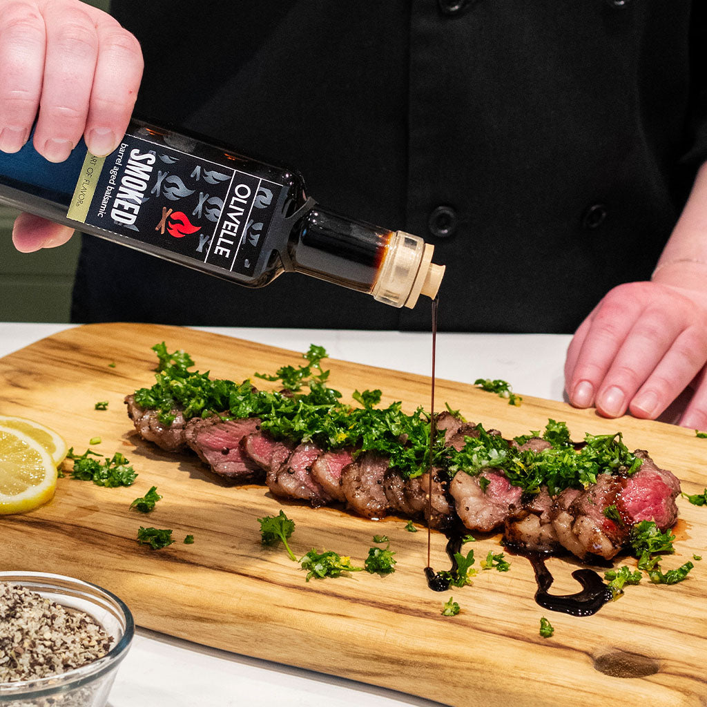 Person pouring sauce over a steak with herbs on a wooden cutting board.