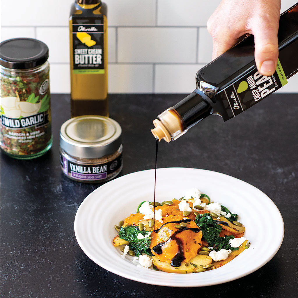 Person pouring olive oil over a dish on a kitchen counter with various ingredients in the background.