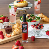 Bottle of Olivelle strawberry jam on a table with strawberries, croissants, and a salad.