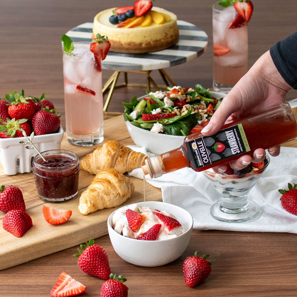 Person pouring syrup into a glass with a dessert, surrounded by strawberries, croissants, and a salad.