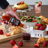 Hand pouring syrup from a bottle labeled 'Stray' into a jar, with a variety of food items including strawberries, croissants, and a salad in the background.