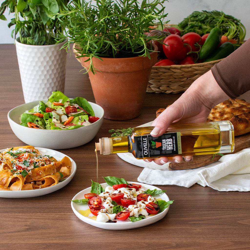 Person pouring olive oil over a salad on a wooden table with various dishes and plants in the background.