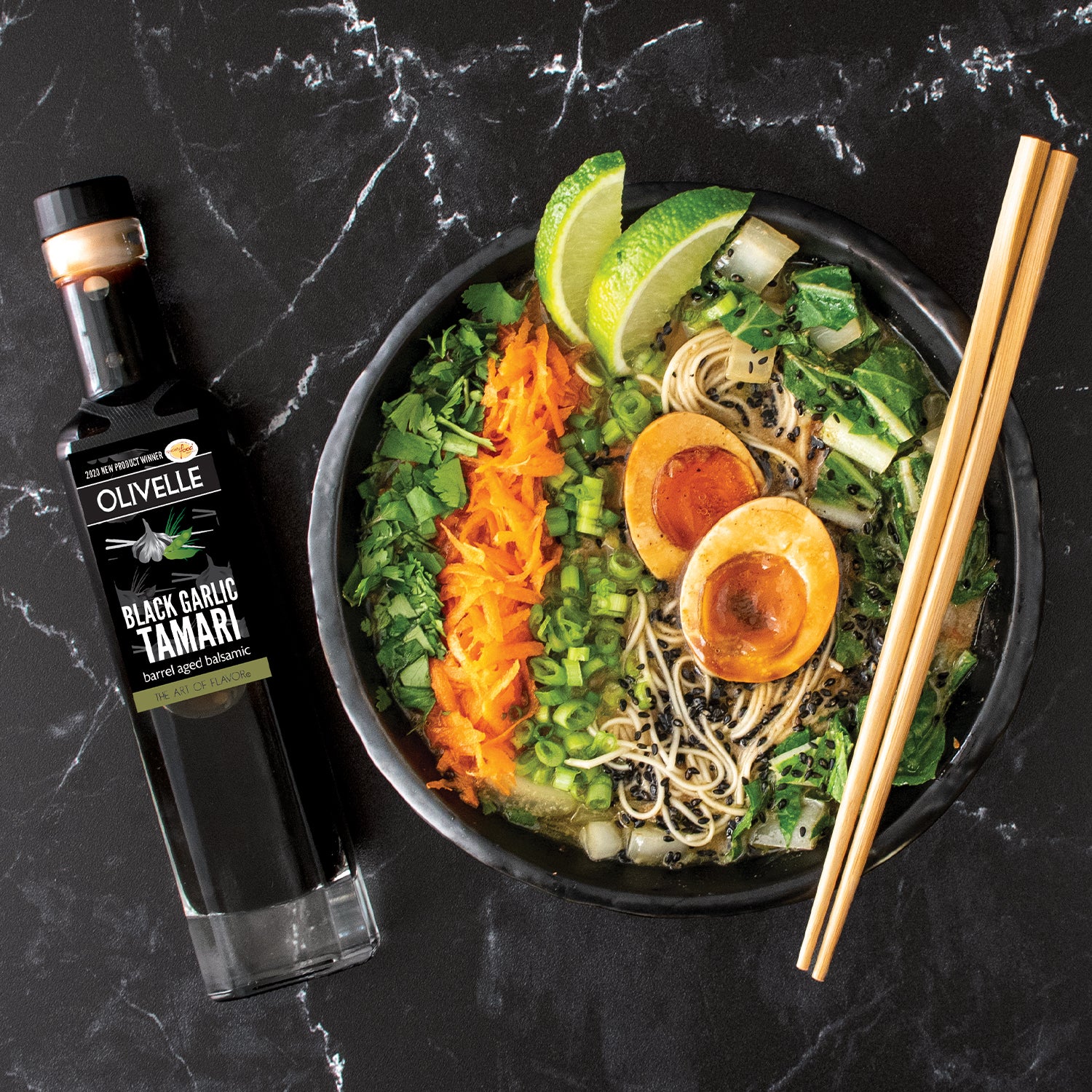 Bowl of ramen with garnishes and a bottle of Olivelle Black Garlic Tamari on a dark marble surface.