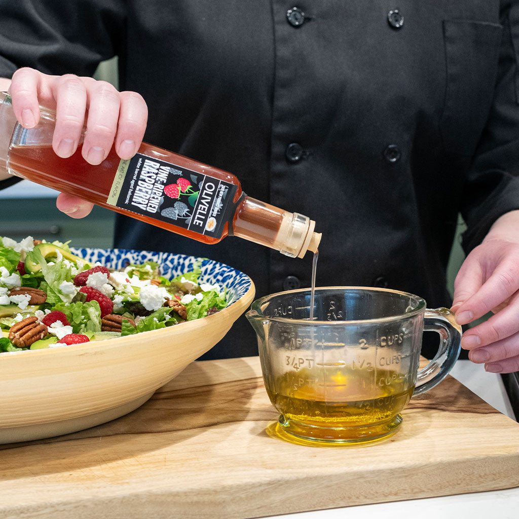 Person pouring a dressing from a bottle into a measuring cup on a wooden cutting board with a salad.