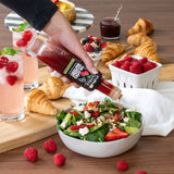 Person pouring a dressing over a salad with croissants and berries on a wooden table.