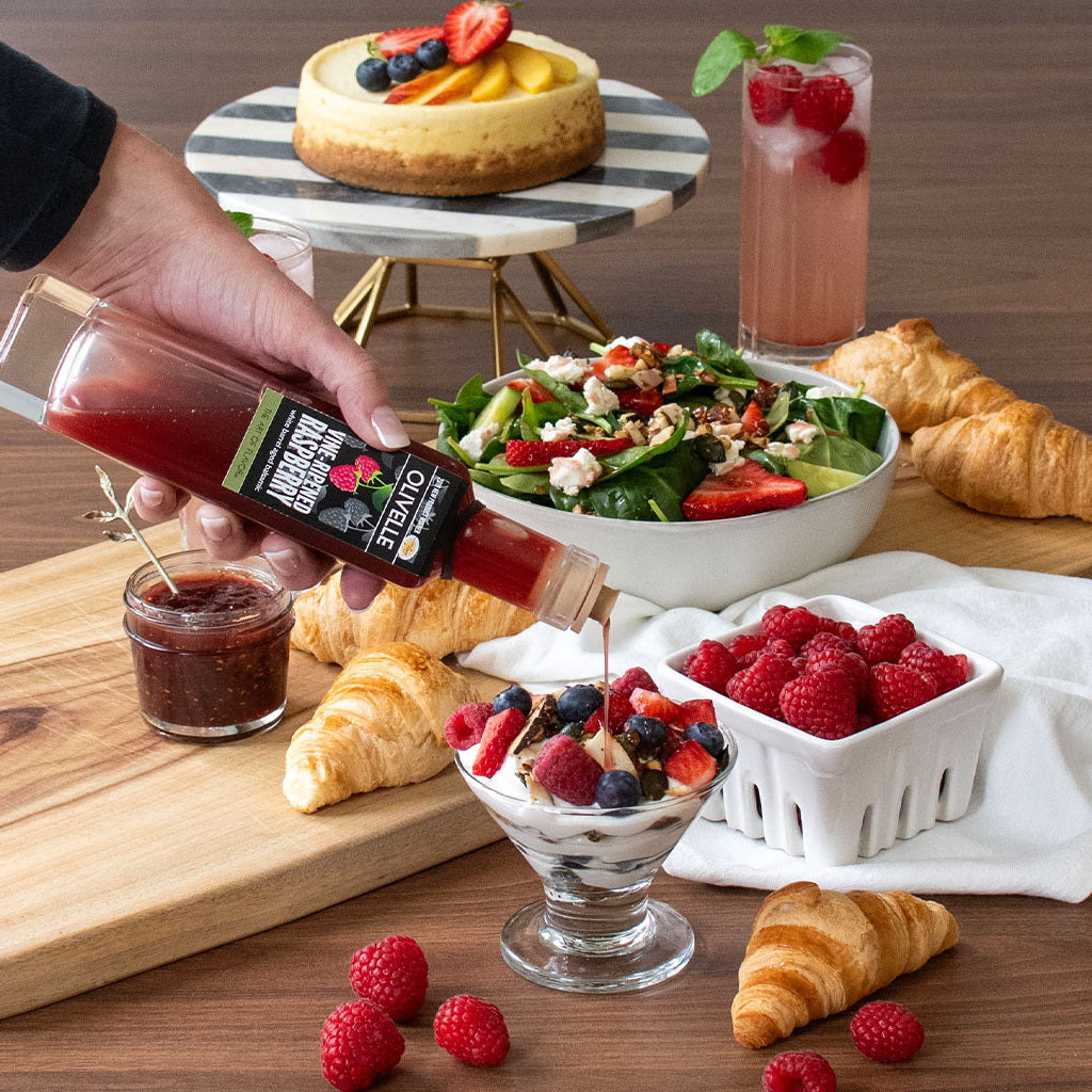 Hand pouring a bottle of syrup over a dessert with a salad, croissants, and berries on a wooden table.