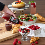 Hand pouring a bottle of syrup over a dessert with a salad, croissants, and berries on a wooden table.