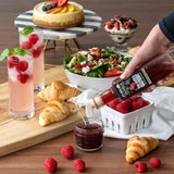 Table setting with drinks, pastries, salad, and a person pouring syrup over raspberries.