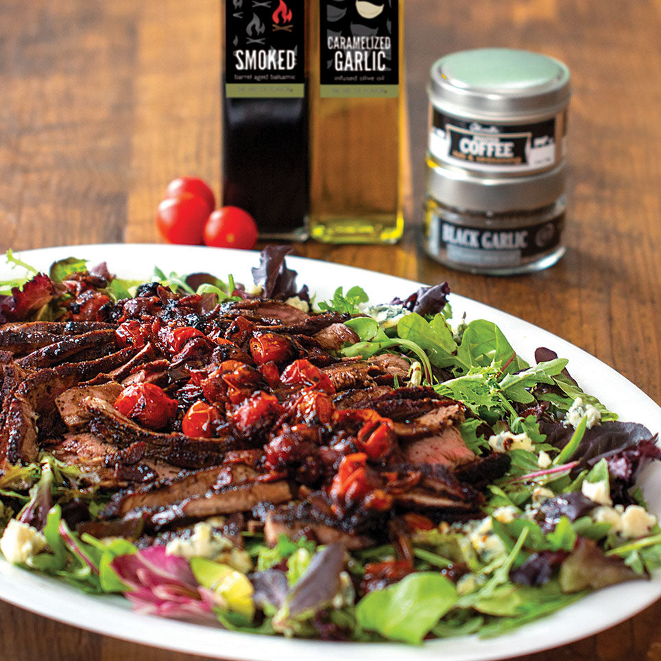 Plated dish of grilled meat with salad on a wooden table with coffee and olive oil bottles in the background.