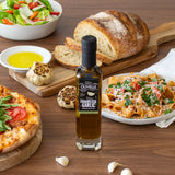 Bottle of Olvelle Caramelized Garlic infused olive oil on a wooden table with pasta, bread, and salad.