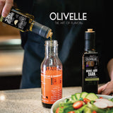 Person pouring olive oil from Olivelle bottle into a glass bottle with salad in the foreground.