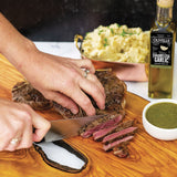 Person cutting a steak on a wooden cutting board with a bottle of Olivelle caramelized garlic oil in the background.
