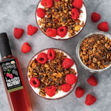 Two bowls of yogurt with granola and raspberries, a bottle of Olivelle raspberry vinegar, and additional raspberries on a gray surface.