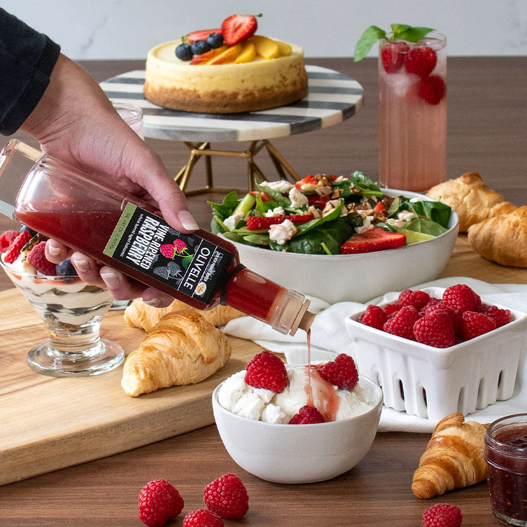 Person pouring raspberry sauce into a dessert with a table full of food including salad, cheesecake, and croissants.