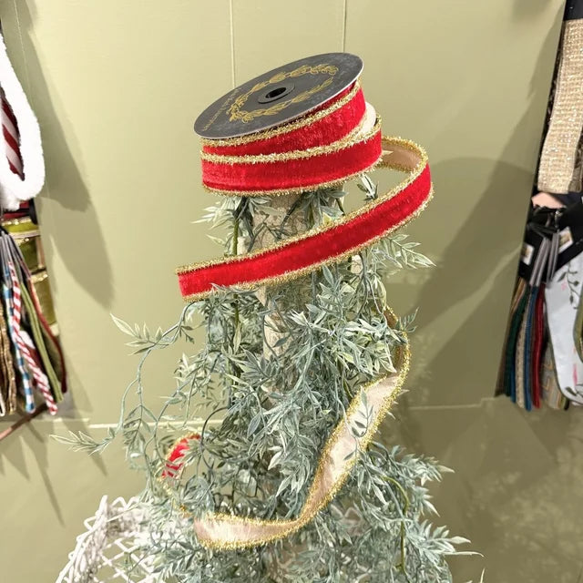 A spool of red velvet ribbon with gold trim wrapped around a frosted green artificial tree in a store display.