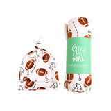A football-themed swaddle blanket shown in two forms: one flat and tied, displaying the white fabric with brown footballs and black tactical play icons, and one rolled with a mint green paper band that reads 'Gigi and Max SWADDLE BLANKET'.