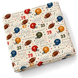 A folded fabric item, likely a quilt or blanket, featuring an American football themed pattern on a cream background with red, blue, orange, and brown helmets, footballs, numbers, and stars.