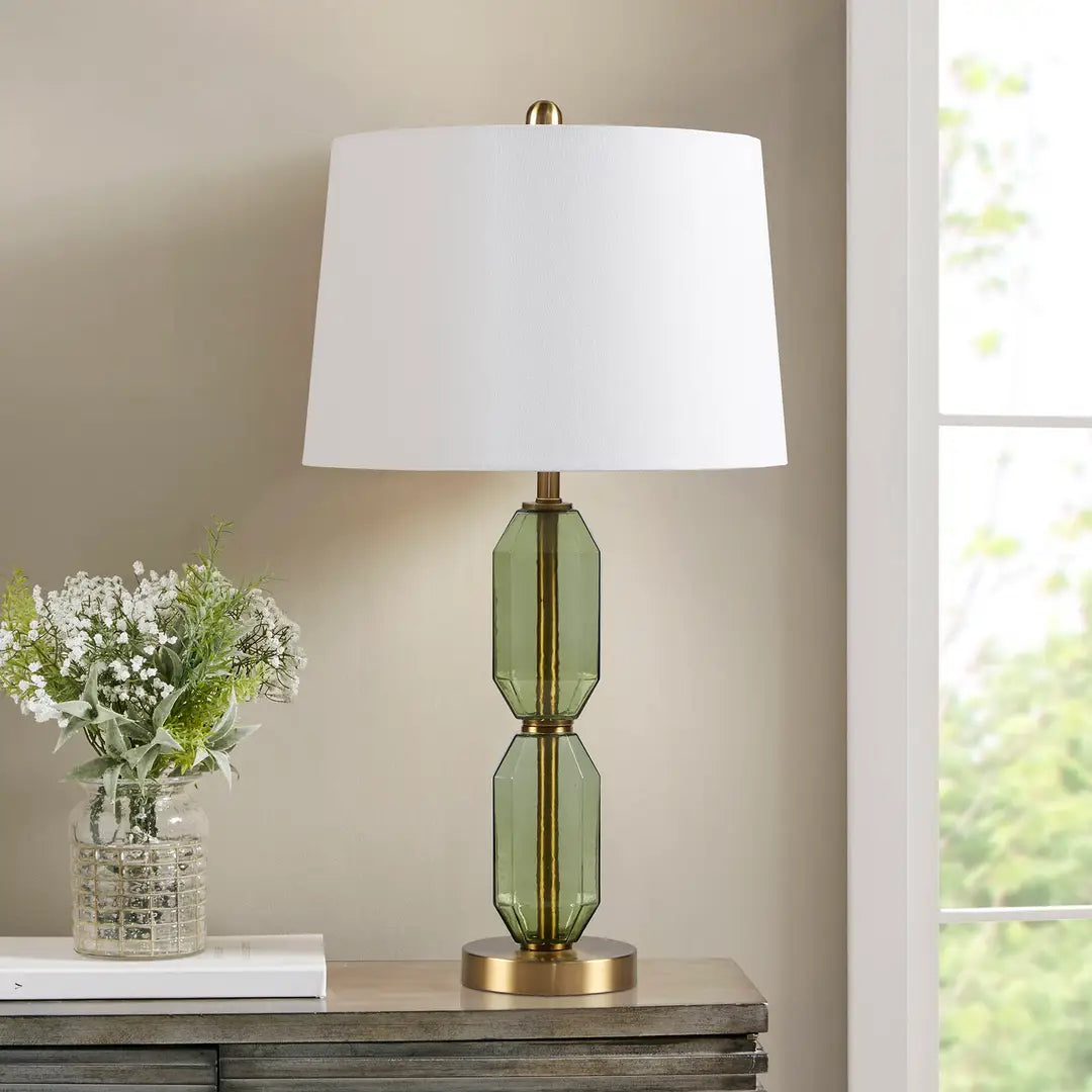 Table lamp with green glass base and white shade on a wooden surface next to a vase of flowers.