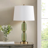 Table lamp with green glass base and white shade on a wooden surface next to a vase of flowers.
