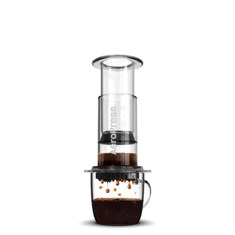 A clear AEROPRESS coffee press with coffee grounds and a cup underneath, showcasing the brewing process.