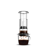 A clear AEROPRESS coffee press with coffee grounds and a cup underneath, showcasing the brewing process.