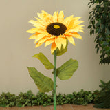 A large artificial sunflower with yellow petals and a brown center, standing upright with green leaves.