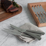 Steak knifes on napkin other steak knifes in the storage box piece of meat on cutting board that has been cut.  All on a marble counter top 