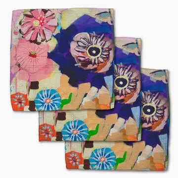 Three folded square napkins with an abstract floral and eye-like pattern in pink, blue, purple, orange, green, and beige, arranged overlapping on a white background.