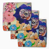 Three folded square napkins with an abstract floral and eye-like pattern in pink, blue, purple, orange, green, and beige, arranged overlapping on a white background.