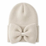 A cream-colored ribbed knit beanie hat featuring a large matching fabric bow on the front, secured with a small pearl-like center bead, shown against a plain white background.