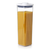 A clear OXO POP container with a small square shape, filled with yellow spaghetti noodles. It has a white lid with a push-button airtight seal.
