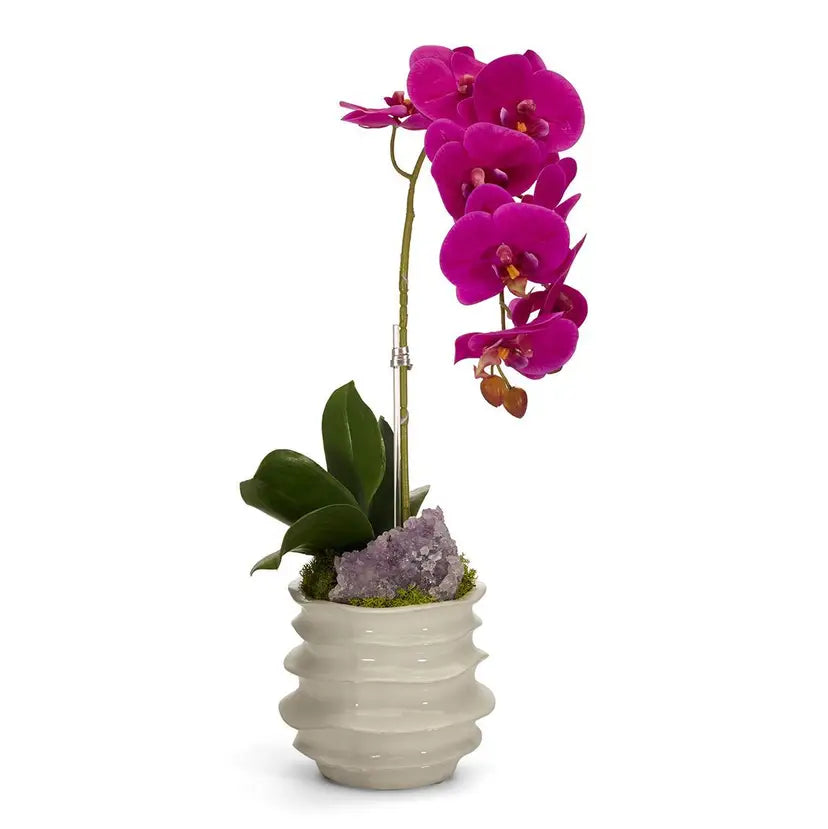 A vibrant magenta Phalaenopsis orchid flower in a textured white ceramic pot with green leaves and a purple crystal accent, set against a plain white background.