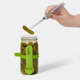 Hand using a pickle-lifting tool to remove a pickle from a jar on a white background