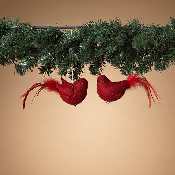 Two red cardinal bird ornaments with feather details, hanging from greenery against a beige background.