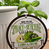Packaging label for a Spinach & Artichoke baked dip mix with the brand Lambs & Thyme displayed, set against a background featuring a white cup and green herbs.