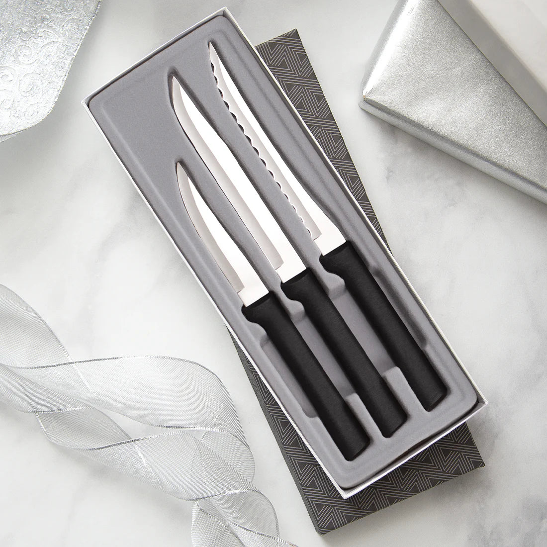 A three-piece kitchen knife set with black handles and silver stainless steel blades, presented in an open gray gift box on a white marble surface with decorative ribbon and a silver box in the background.